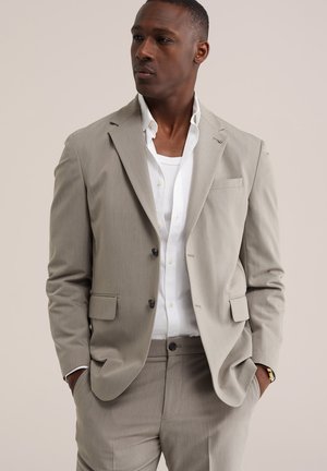 Man draagt een beige colbert en broek met een wit overhemd en wit T-shirt eronder, handen in de zakken, kijkt naar links.