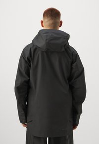 Czarna wodoodporna kurtka z kapturem, o smukłym designie i gładkiej fakturze. Zawiera oznaczenie GORE-TEX na dolnej części pleców.
