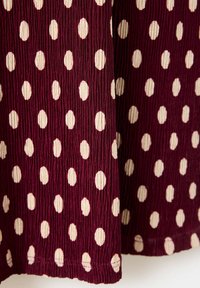 Tissu bordeaux texturé avec un motif de pois crème irrégulier et des coutures visibles sur le bord inférieur.