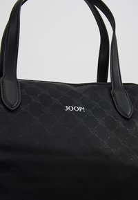 Sac à main noir en tissu à motif subtil, poignées en cuir, fermeture éclair et logo argenté "JOOP!" sur le panneau avant central.