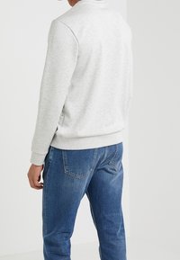 Mann in hellgrauem langärmeligen Sweatshirt und blauen Jeans, stehend mit dem Rücken zur Kamera vor einem schlichten weißen Hintergrund.