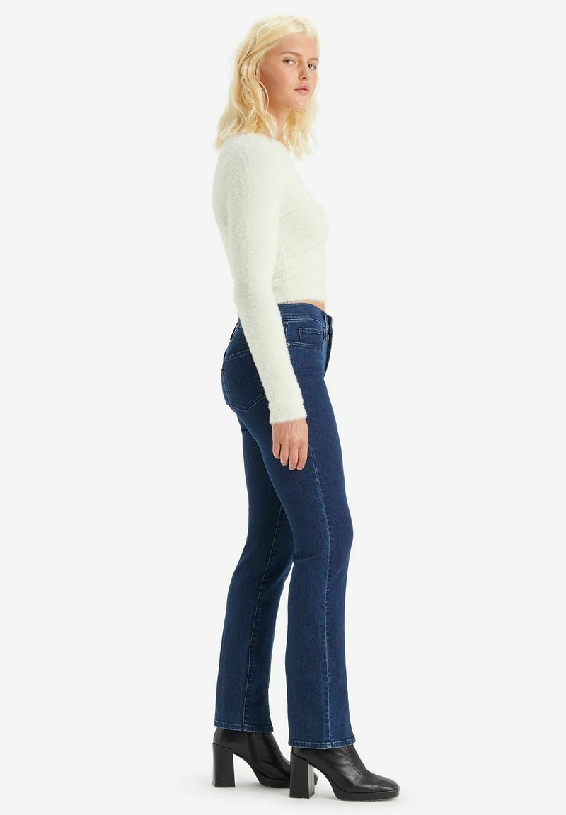 Donna bionda che indossa un top corto a maniche lunghe bianco e peloso, jeans a vita alta blu scuro e stivaletti neri con tacco, in posizione laterale.