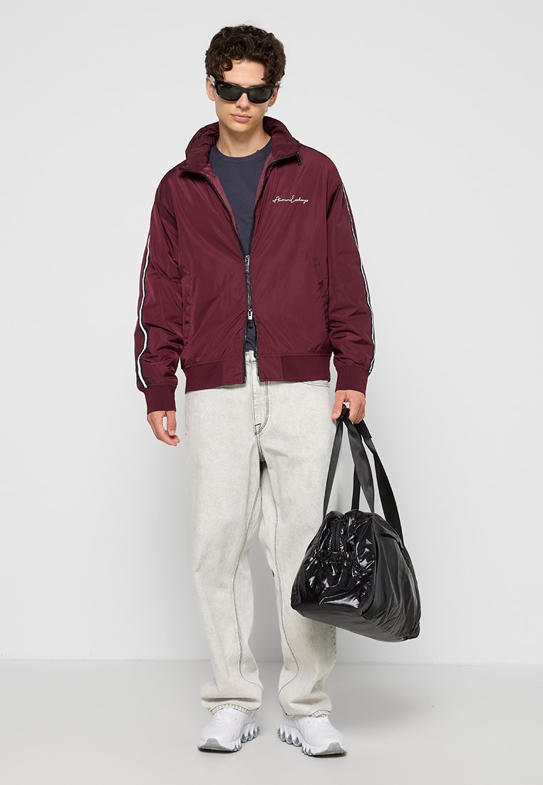 Giacca bomber bordeaux con strisce bianche sui lati, materiale leggero, indossata sopra una maglietta blu navy. Jeans grigi chiari e una borsa a tracolla nera lucida.