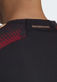 Mustan urheilupaita, jossa on teksturoitu kangas, punaiset yksityiskohdat olkapäissä ja kultainen "DEUTSCHLAND" -merkki takakanavalla.