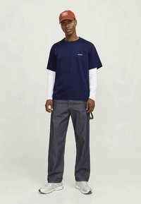 Camiseta azul-marinho sobre uma camisa de mangas longas branca, combinada com calças de ganga escuras e ténis cinzentos. Boné vermelho com detalhes de logótipo.