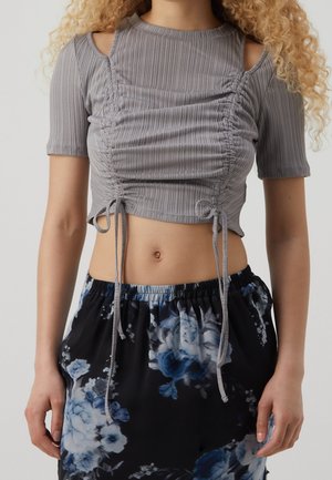 Kvinde iført en grå rynket crop top med skulderudskæringer og et sort skørt med blå og hvide blomsterprint.