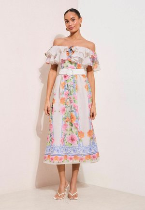 Kobieta w białej midi sukience z odkrytymi ramionami, falbanami, kwiatowym wzorem, białym paskiem, złotymi kolczykami kołami i białymi sandałami na obcasie, stojąca przy ścianie.