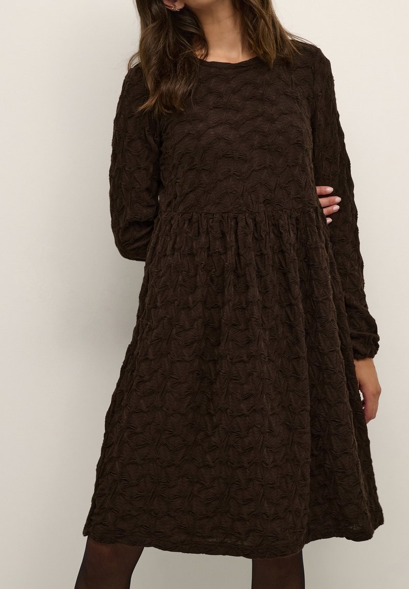 Femme portant une robe texturée à manches longues de couleur brun foncé avec une taille froncée et une jupe ample et fluide sur un fond neutre.