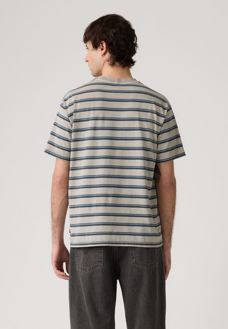 Man facing away wearing light gray short-sleeve shirt with dark horizontal stripes and dark gray jeans against plain background.