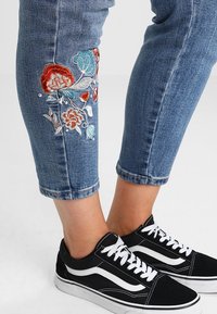 Modré džíny z denimu, zkrácené s vyšívanými květinovými detaily v červené, modré a bílé barvě, spárované s černými teniskami s bílými akcenty.