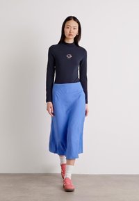 Svart langermet topp med logo, kombinert med et blått midi-skjørt. Modellen har på seg rosa joggesko og hvite sokker, med en jevn tekstur generelt.