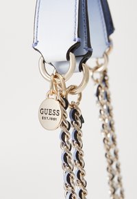 Cinturino in ecopelle azzurro chiaro con hardware dorato, caratterizzato da un design a catena e un'etichetta circolare impressa con "GUESS EST.1981."