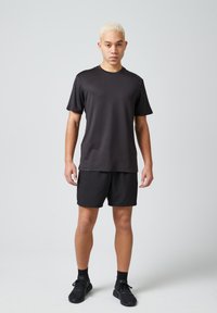 Czarny sportowy T-shirt z krótkimi rękawami, w zestawieniu z czarnymi szortami. Materiał wydaje się lekki i gładki. Noszony z czarnymi butami sportowymi.