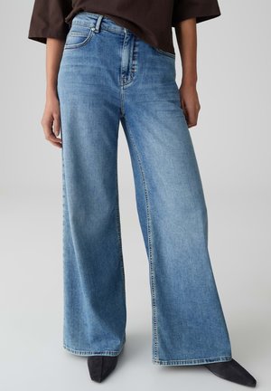 Person, die hellblaue Jeans mit weitem Bein und schwarze spitze Schuhe trägt, steht vor einem einfarbigen grauen Hintergrund.