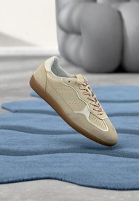 Beige Leder-Sneaker mit einer hellgrauen Zunge, strukturierten Paneelen, rosafarbenen Schnürsenkeln und einer Gummisohle, vor einem blau strukturierten Teppich.