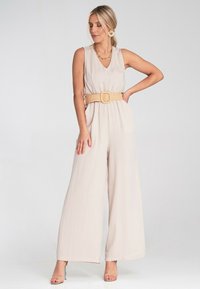 Beżowy kombinezon o szerokich nogawkach, dekolt w kształcie litery V, zwężona talia za pomocą plecionej paska, gładka tkanina i minimalistyczna estetyka.