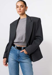 Tmavě šedý strukturovaný blazer přes světlý šed�ý svetr, doplněný modrými džíny a černým opaskem. Blazer má široké klopy a volný střih.