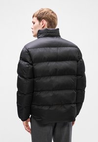 Μαύρο μπουφάν πούφ με ψηλό γιακά, που διαθέτει ραμμένα τμήματα και λεία, γυαλιστερή υφή. Χωρίς ορατό υλικό ή διακοσμήσεις.