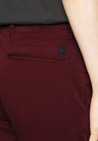 Burgundské kalhoty s hladkou texturou, mají zadní kapsu s černou etiketou a kontrastním prošíváním. Viditelný slim fit střih.