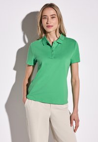 Grön kortärmad polo med krage och treknappslås, i kombination med ljusa, löst sittande byxor. Slät tygtextur.