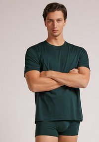 Ciemnozielona koszulka z krótkim rękawem o okrągłym dekolcie i gładkiej fakturze, zestawiona z dopasowanymi krótkimi spodniami. Prosty, nowoczesny design.