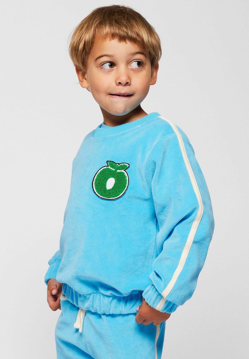 Svijetloplava flisna majica s kapuljačom, s zelenim vezenim motivom voća, bijelim bočnim prugama i elastičnim pojasom. Meka tekstura i opušteni kroj.