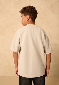 Camicia grigia chiara a maniche corte con motivo strutturato a griglia, scollo arrotondato e vestibilità rilassata, abbinata a pantaloni in denim scuro.