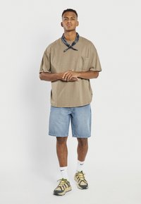 Bărbat tânăr stând cu mâinile împreunate, purtând un tricou bej supradimensionat, pantaloni scurți din denim albastru deschis, șosete albe Nike și adidași bej cu șireturi verzi.