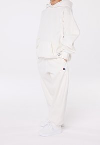 Barn som bär en vit hoodie, matchande svettbyxor med logotyp och vita sneakers, står med händerna i fickorna på vit bakgrund.