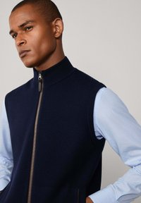 Mornarsko pleteno bezrukavno pletivo s visokim ovratnikom i punim zatvaračem, u kombinaciji s svijetloplavom košuljom dugih rukava ispod.