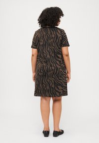 A person with curly hair stands facing away, wearing a short brown dress with a tiger stripe pattern and black shoes.