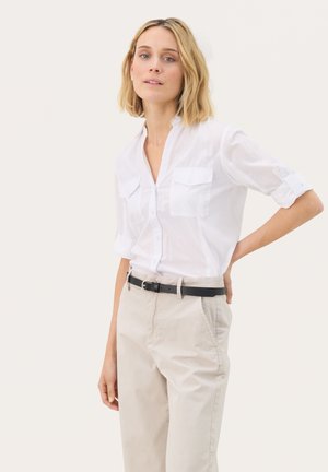 Camicia bianca leggera con bottoni, maniche arrotolabili e due tasche sul petto, abbinata a pantaloni beige e una cintura nera. Texture liscia.