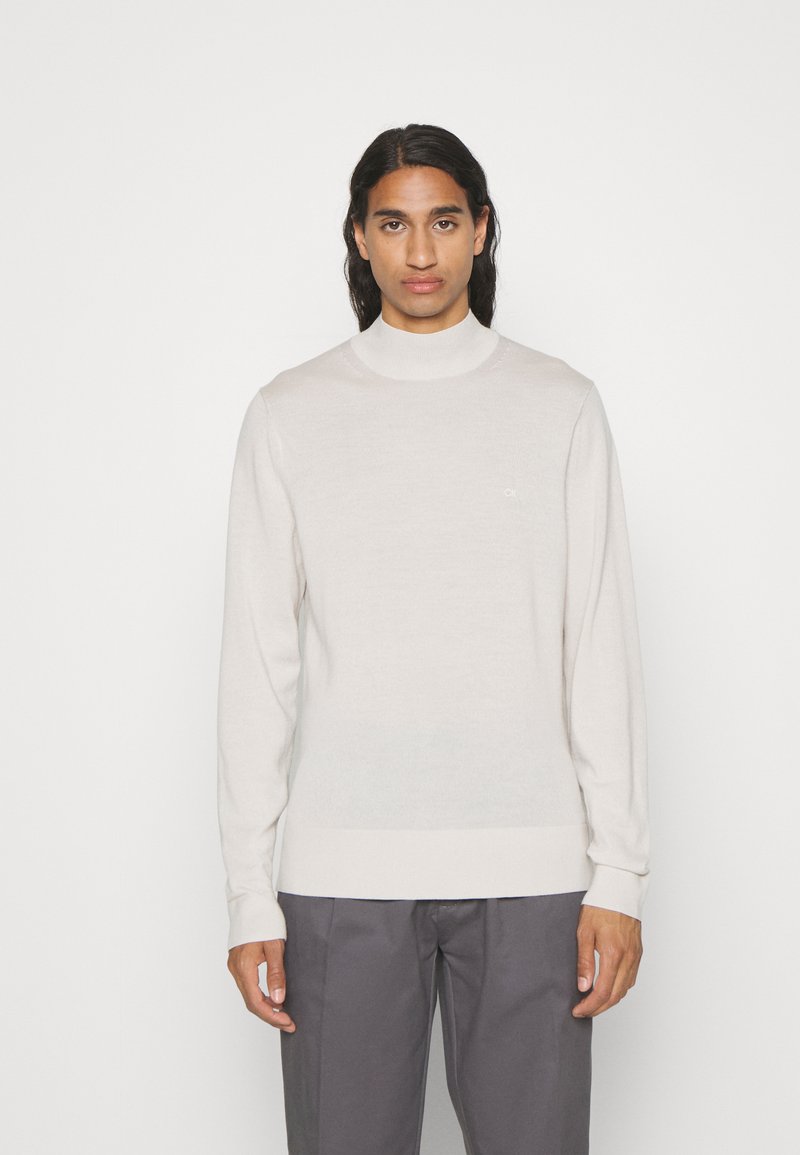 Maglione a collo alto beige chiaro in un tessuto morbido, con maniche lunghe e un discreto logo sul petto. Design aderente con polsini a coste.