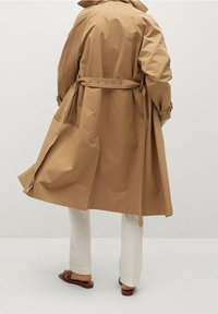 Trench-coat beige avec taille ceinturée, comportant des épaulettes et des fentes latérales, porté sur un pantalon blanc et des sandales marron. Texture lisse.