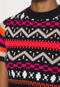 Neulottu neulepaita, jossa on laivastonsinisiä, vaaleanpunaisia, valkoisia ja oransseja geometrisia kuvioita, lyhyet hihat ja resorinen pyöreä kaulus.