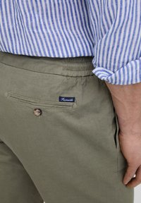 Los pantalones en un suave tono verde oliva cuentan con una cinturilla elástica y un bolsillo trasero con cierre de botón. Se puede ver una pequeña etiqueta de la marca en azul oscuro.