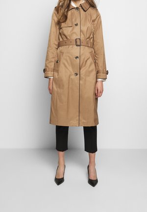 Kvinde iført en beige trenchcoat med bælte og knapdetaljer, sorte kapribukser og sorte spidse hæle, stående mod en ensfarvet baggrund.