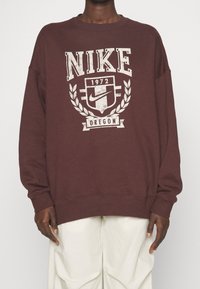 Vinröd Nike-tröja med oversized passform, med en sliten vit logotyp och skölddesign, texten "1972" och "Oregon", ribbade muddar och fåll.