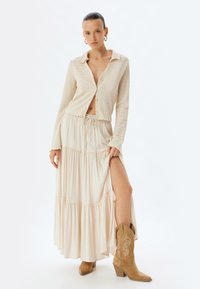 Chemise à boutons beige texturée avec un décolleté en V profond, associée à une jupe maxi crème fluide dotée de couches superposées et d'une fente sur le côté, portée avec des bottes beige.