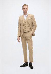 Costum bej format trei piese, cu o jachetă ajustată, vestă și pantaloni evazați. Asortat cu o cămașă albă și pantofi din piele neagră.
