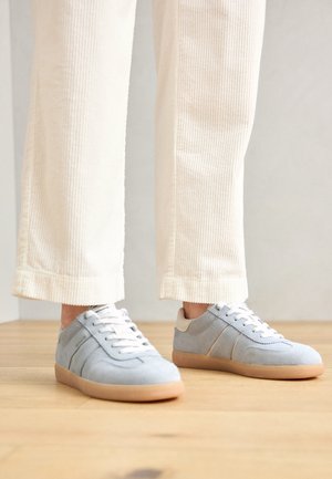 Nogi w kremowych, prążkowanych spodniach i jasnoniebieskich zamszowych trampkach ze sznurowadłami w kolorze białym i gumowymi podeszwami, stojące na drewnianej podłodze.