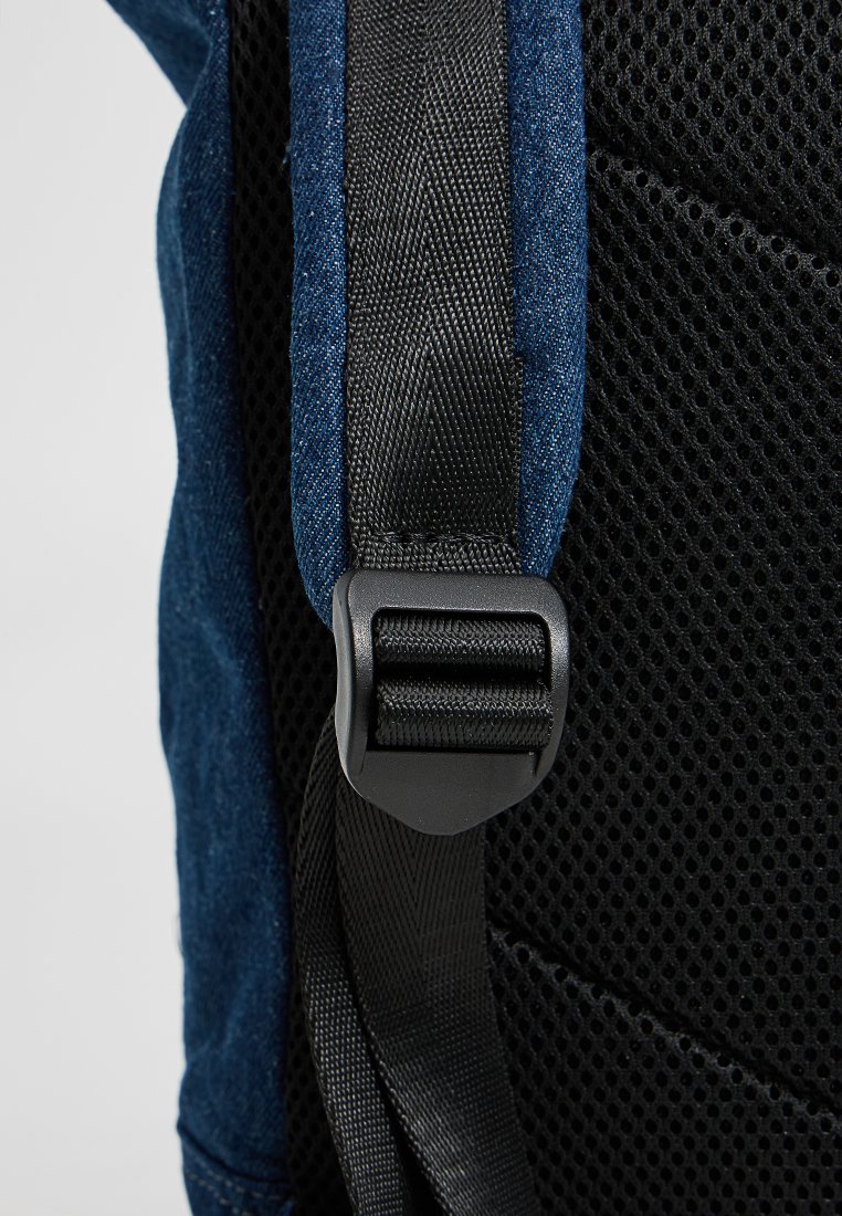 Detail van een donkerblauwe stoffen rugzak, met een textuurpanneel van zwart gaas en een verstelbare zwarte strap met een plastic gesp.
