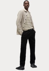 Beige struktureret yderjakke med sorte knapper over en cremefarvet stribet sweater, parret med sorte corduroy-bukser og glatte sorte sko.