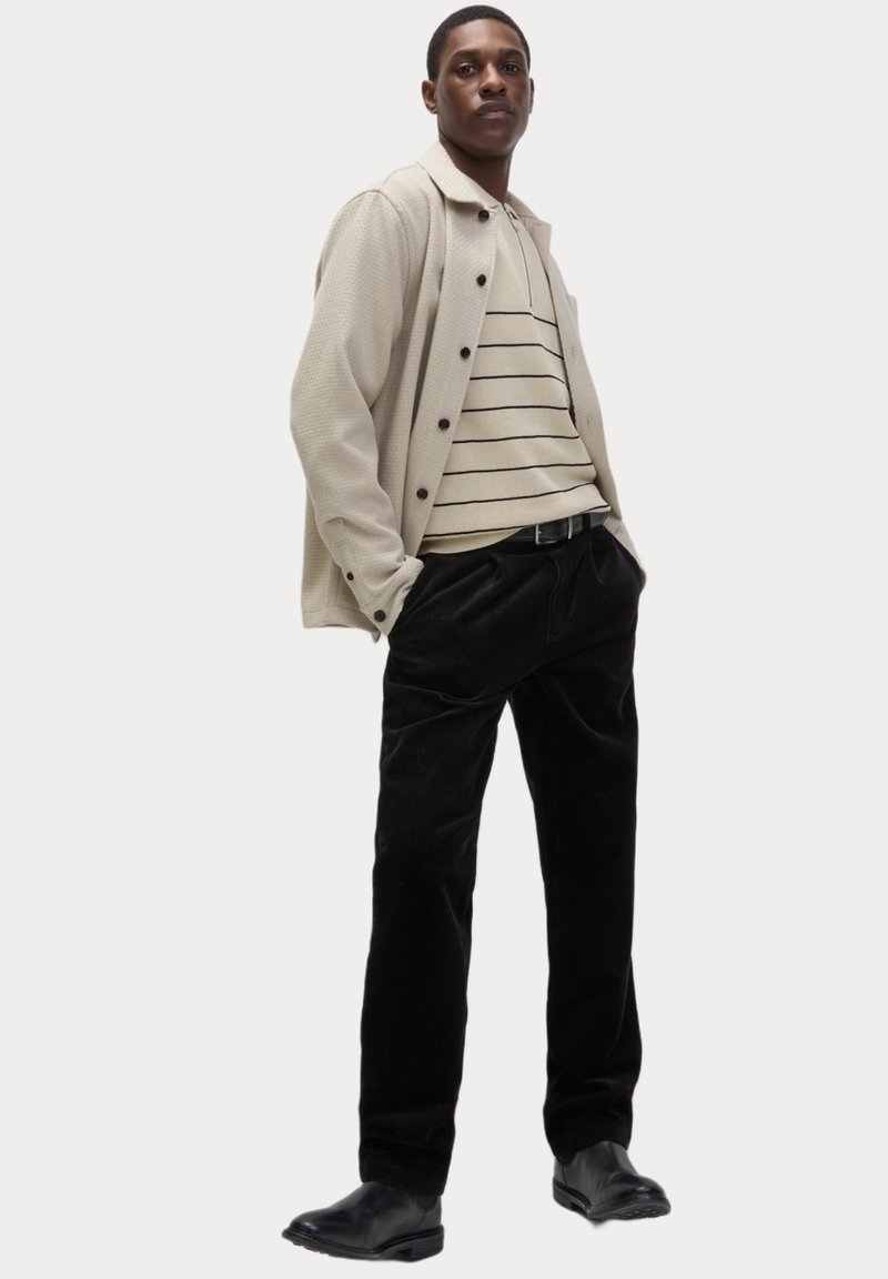 Beige struktureret yderjakke med sorte knapper over en cremefarvet stribet sweater, parret med sorte corduroy-bukser og glatte sorte sko.