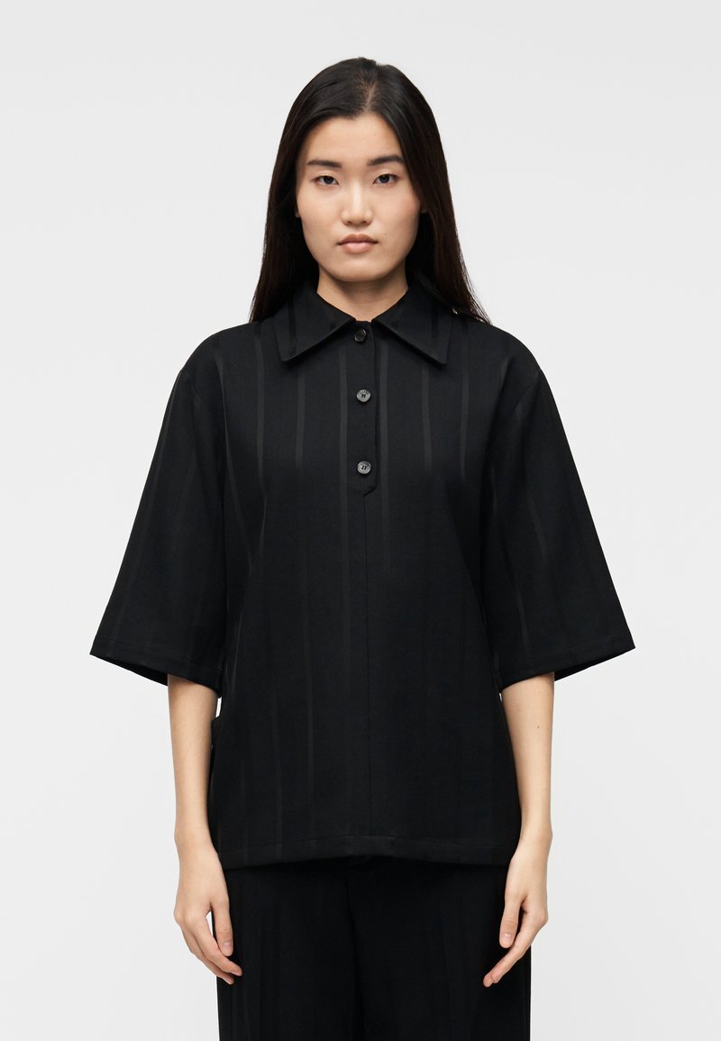 Camicia nera con colletto e maniche corte, caratterizzata da righe verticali e tre bottoni sul davanti. Il tessuto sembra liscio e leggero.