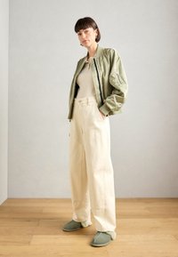 Olijfgroene bomberjack, crèmekleurig ribbed top, wijde beige broek met zakken en zachte groene slippers met bontaccenten. Houten vloer achtergrond.