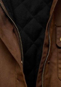 Brun jakke med en gylden lynlås, med en quiltet sort foring synlig indeni. Yderligere detaljer inkluderer inderlommer og en robust tekstur.