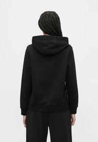 Čierna mikina s kapucňou so sťahovacou šnúrkou, mäkkou textúrou, rebrovanými manžetami a lemom. Navrhnutá s uvoľneným strihom a veľkou, nastaviteľnou kapucňou.