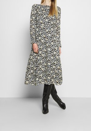 Kobieta ubrana w czarną sukienkę midi w kwiatowy wzór z długim rękawem, w połączeniu z czarnymi skórzanymi kozakami sięgającymi do kolan, stojąca na jednolitej szarej podłodze.