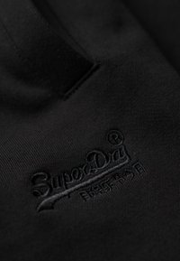 Tessuto nero con una texture liscia, caratterizzato da un logo ricamato in una tonalità più scura. Il design è minimalista, focalizzandosi sul logo.